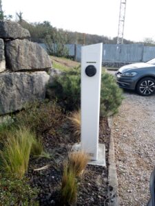 Borne de recharge électrique blanche dans un parking avec une voiture grise à proximité et aménagement paysager.